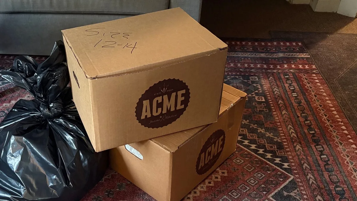 Two cardboard boxes stacked on a patterned rug, the top one marked SIZE 12-14 in marker, next to a tied black donation bag.